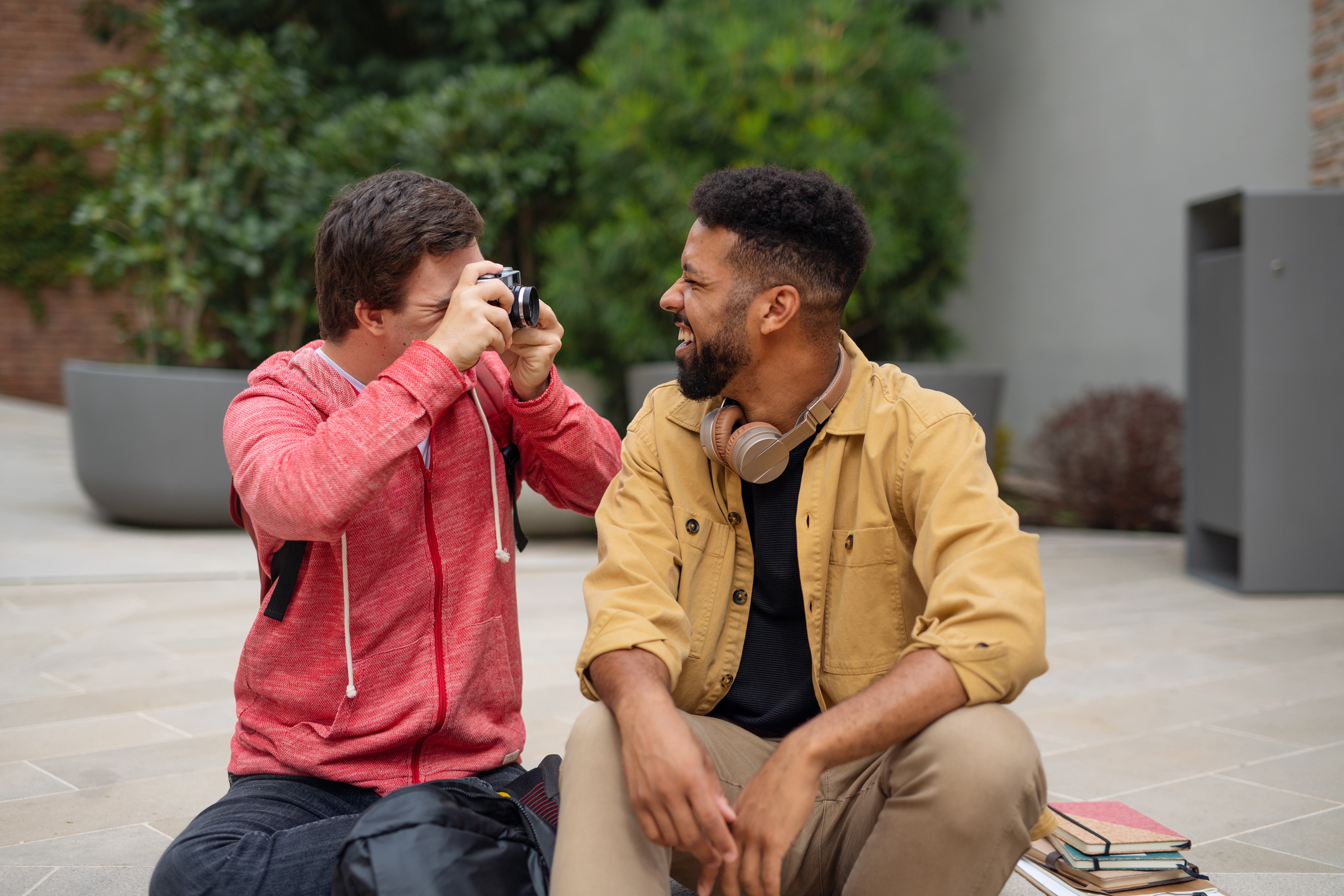 Happy young man with Down syndrome sitting and taking picture with camera of friend outdoors